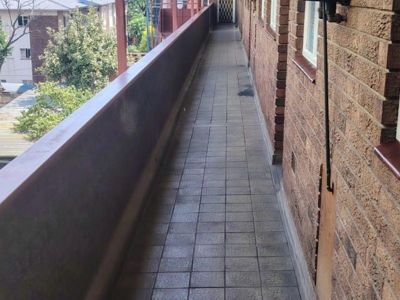 A long, covered exterior balcony featuring brick walls, tiled flooring, and a metal railing, providing access to the apartment entrance.