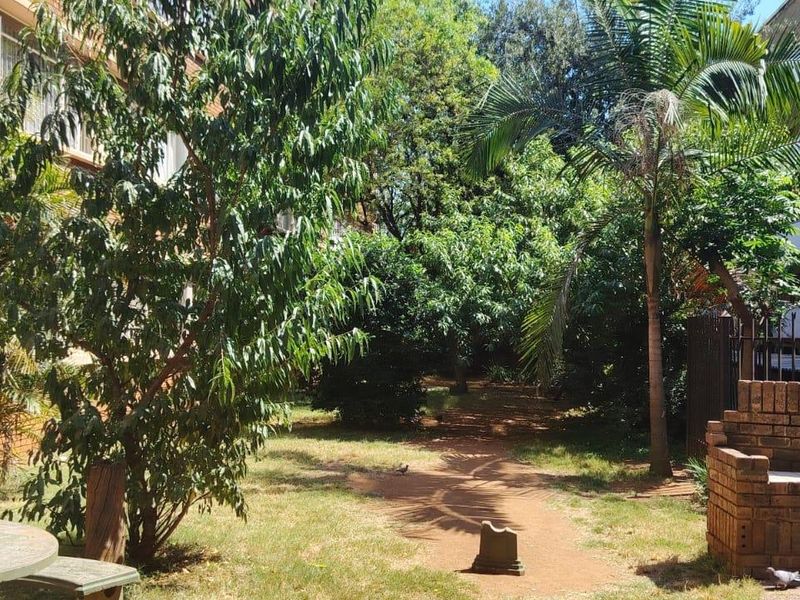 A communal garden area featuring mature trees, a brick built-in braai, and a concrete table set within an apartment complex.