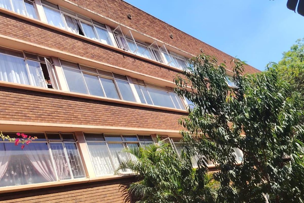 A view of the rear brick facade of a multi-story apartment building featuring large windows and established surrounding greenery.
