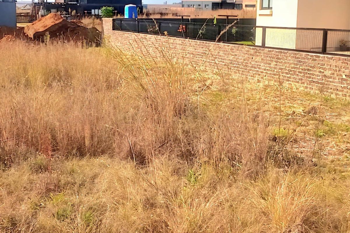 A vacant residential plot featuring overgrown grass and a brick boundary wall with metal fencing.