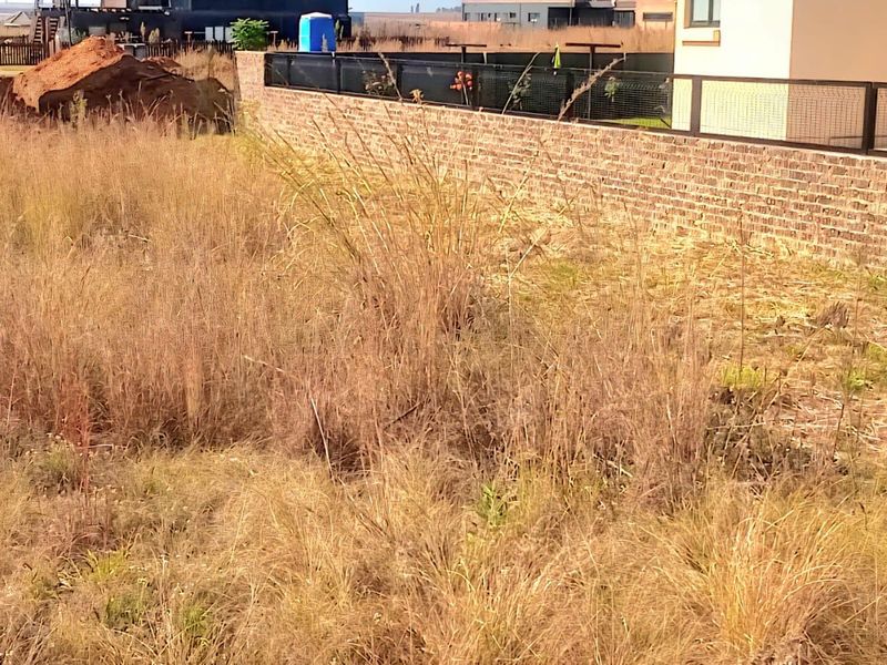 A vacant residential plot featuring overgrown grass and a brick boundary wall with metal fencing.