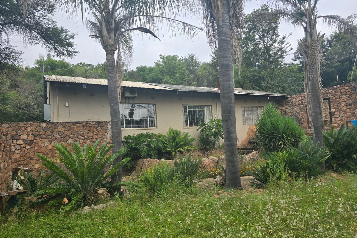 Rear view of a farm-style dwelling featuring stone retaining walls, security-barred windows, and a corrugated metal roof set within a natural, overgrown garden landscape.
