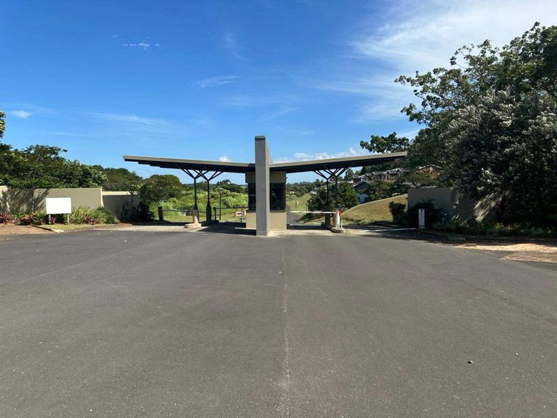 A modern security gatehouse entrance featuring a cantilevered roof structure, boom gates, and paved driveway access to the residential estate.
