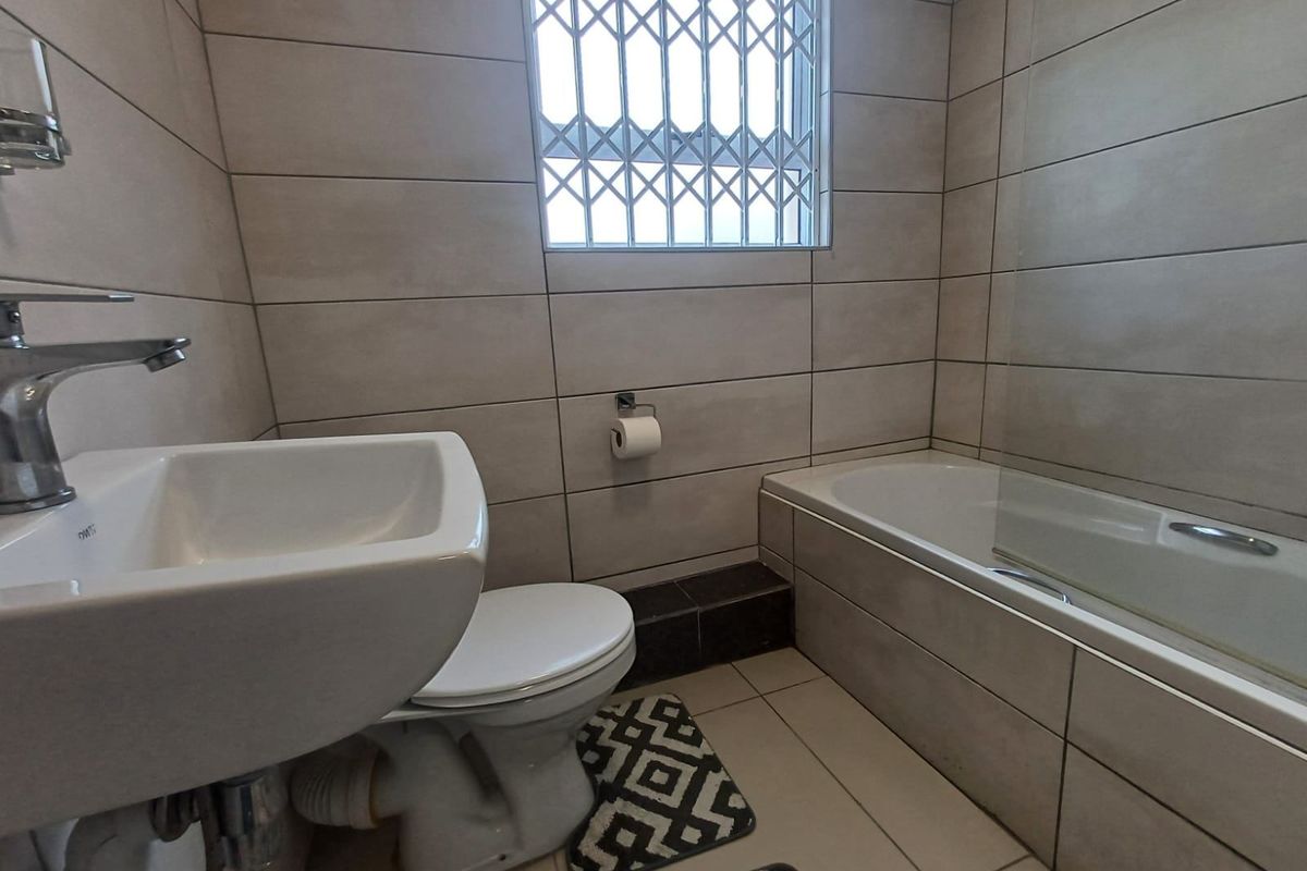A modern bathroom featuring tiled walls and flooring, a wall-mounted basin, a bathtub with a glass screen, and a window with a security gate.