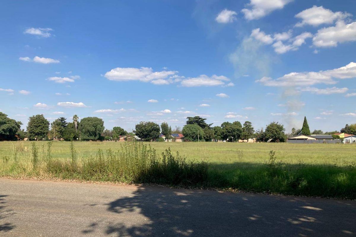 A view of a vacant residential plot from the adjacent asphalt road, featuring open grassland and surrounding mature trees.