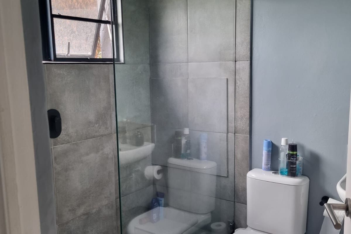 A compact modern bathroom featuring a glass-enclosed shower with a rain shower head, grey tiled finishes, and a standard toilet.