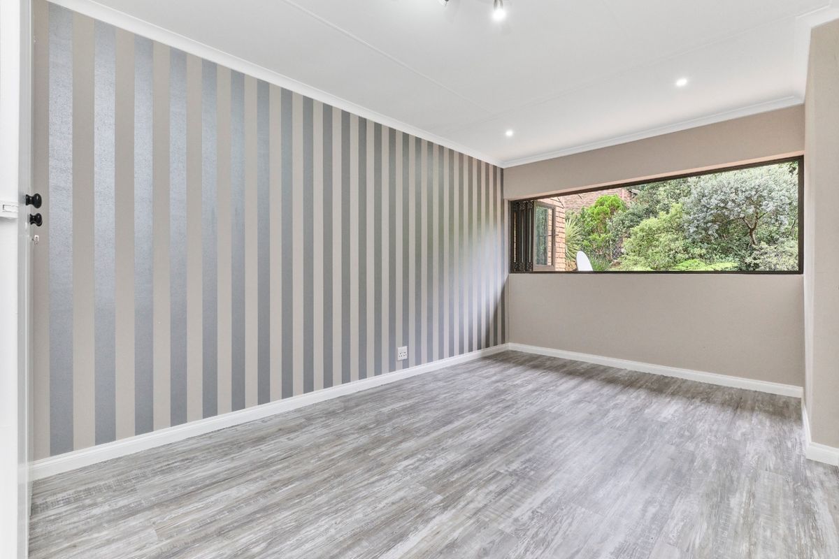 A bright, empty bedroom featuring modern grey laminate flooring, a feature striped wall, and recessed ceiling lighting with a view of the garden.
