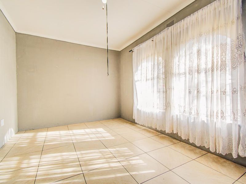 An empty living room featuring neutral tiled flooring, plain walls, and natural light filtering through window curtains.