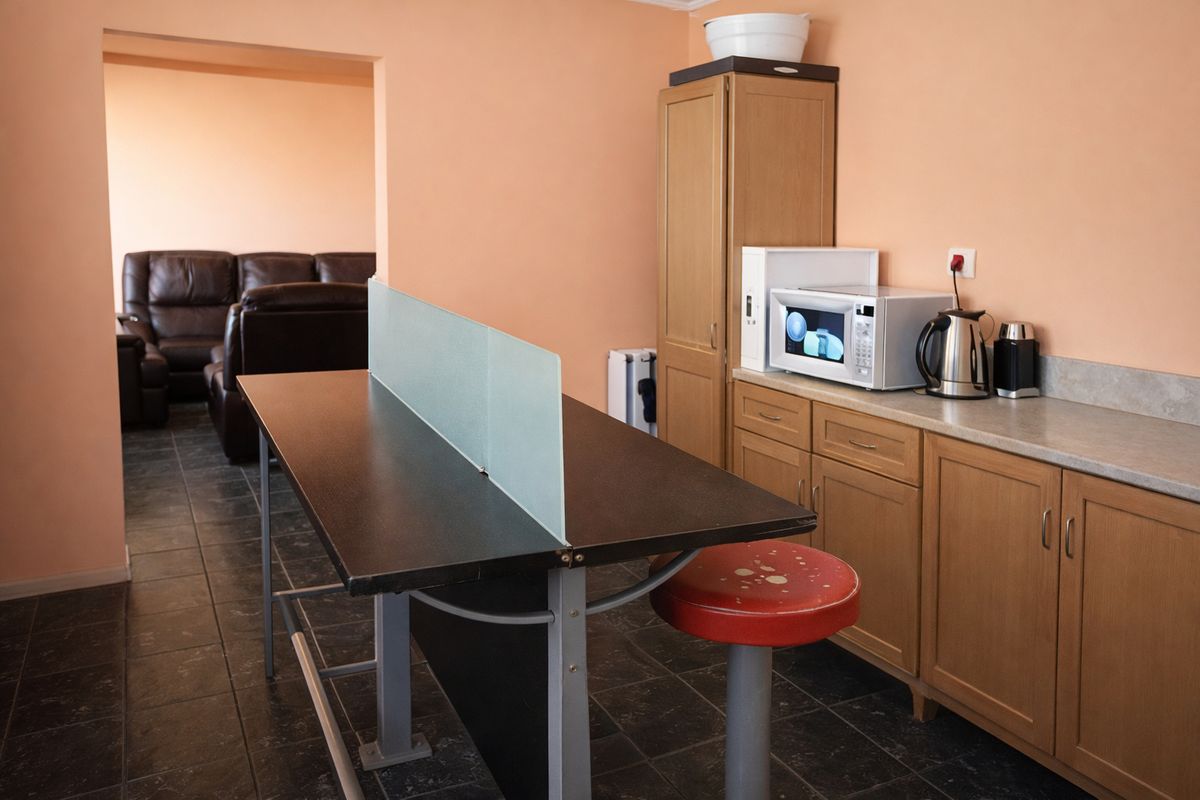 A functional kitchen area featuring built-in wooden cabinetry, laminate countertops, and tiled flooring, with an adjacent living space visible through the doorway.