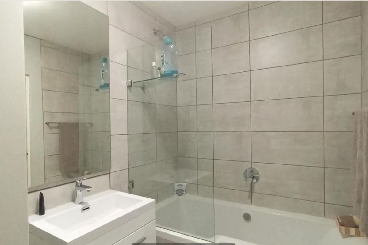 Modern bathroom featuring a vanity with basin, wall-mounted mirror, and a bathtub with a glass shower screen and neutral wall tiling.