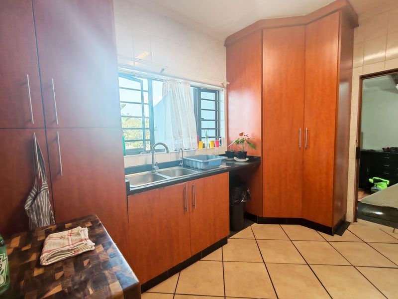A functional kitchen featuring warm-toned built-in cabinetry, a double basin sink, tiled backsplash, and tiled flooring with natural light from the window.