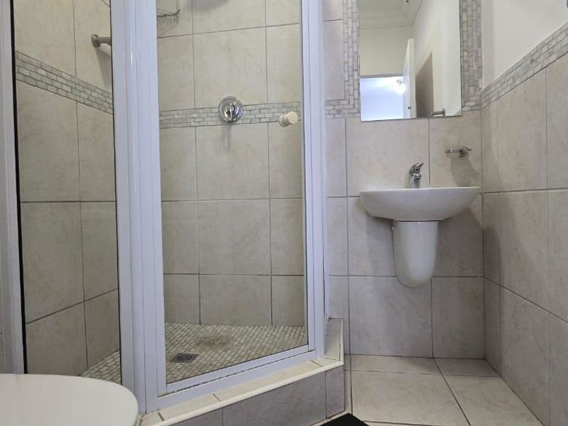A compact bathroom featuring a corner shower enclosure, pedestal basin, and toilet with neutral wall and floor tiling.