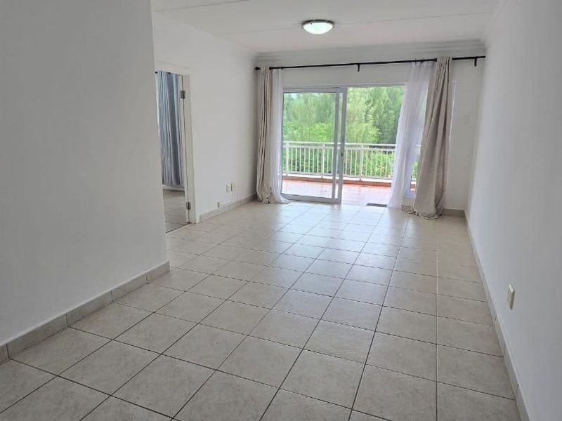 A bright, empty living area featuring neutral tiled flooring and a large sliding glass door providing direct access to an outdoor balcony.