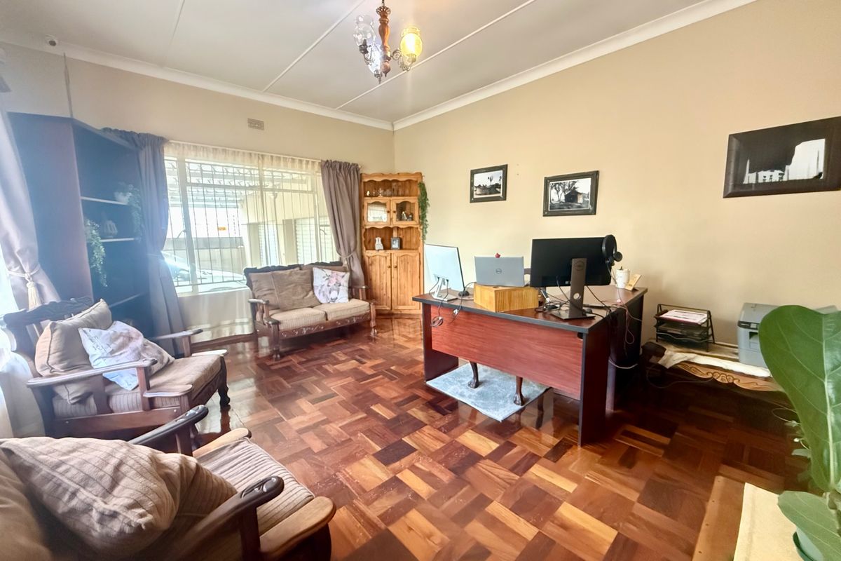 Spacious living area featuring classic parquet flooring, neutral wall tones, and large windows with security bars, currently utilized as a home office space.