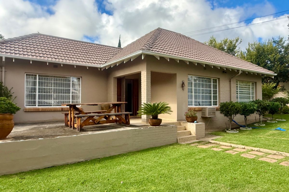 Rear view of a single-story brick house featuring a tiled roof, concrete patio area, and established garden surroundings.