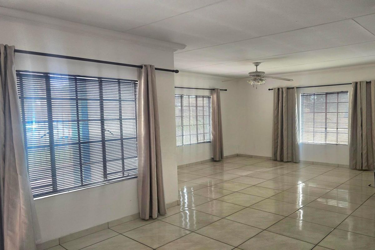 A spacious, empty living area featuring neutral tiled flooring, ceiling fan, and multiple windows fitted with venetian blinds and curtain rods.