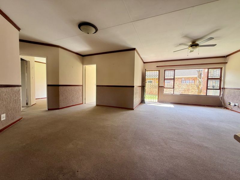 Spacious, empty living room featuring neutral carpeting, ceiling fan, and large windows with security gate access to the exterior.
