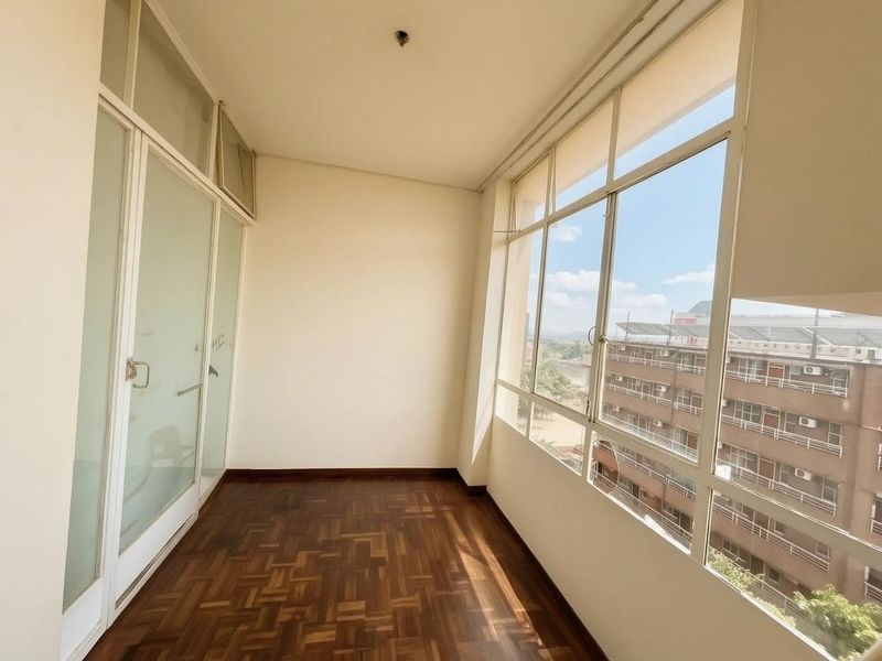 A bright, empty enclosed balcony featuring polished parquet flooring and large windows offering an urban view.