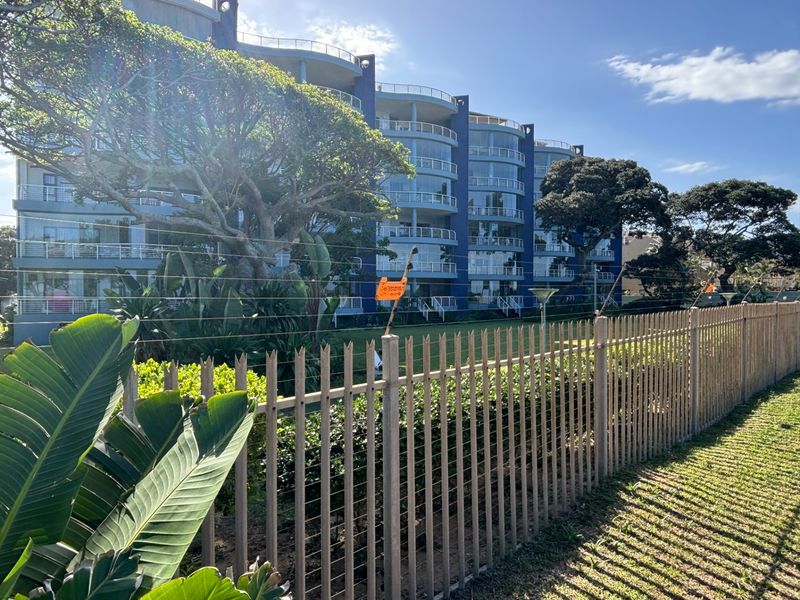 Exterior view of a multi-story apartment building featuring private balconies, landscaped grounds, and secure perimeter fencing.