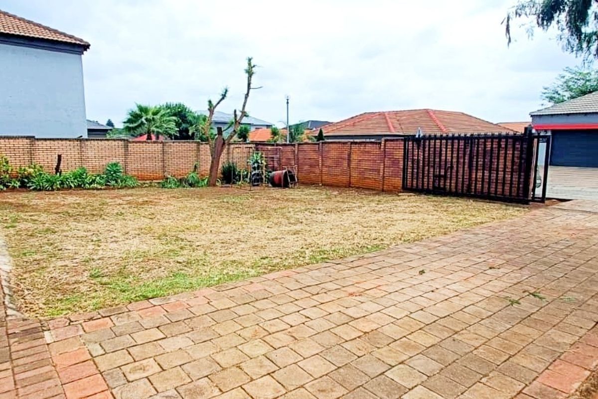 A spacious rear garden featuring a paved patio area, brick boundary walls, and a gated driveway entrance.
