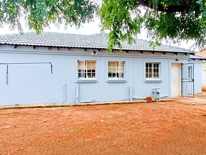 Rear view of the house featuring a tiled roof, security gate, and exterior wall with exposed plumbing, set against an unpaved backyard area.