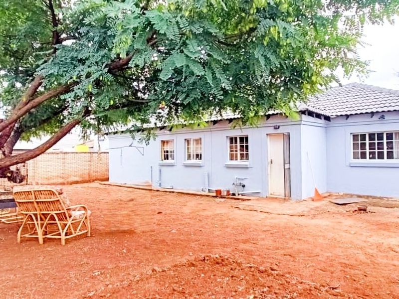 Rear view of a single-story house featuring a large tree, unpaved yard, and exterior access door.