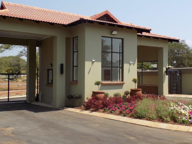 A well-maintained residential estate entrance featuring a tiled roof structure, security gate, paved driveway, and landscaped garden beds.