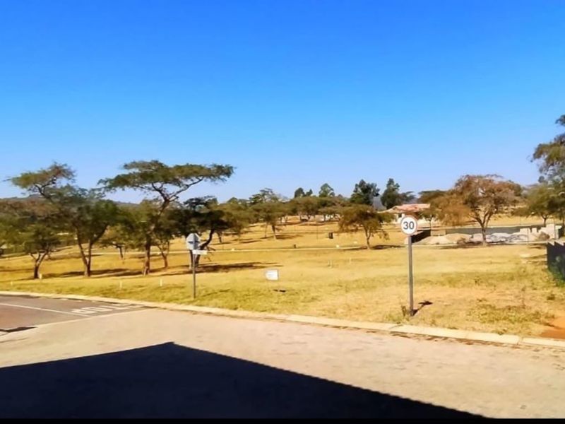 A view of a vacant residential plot featuring a paved driveway, boundary wall, and established trees in a quiet estate setting.