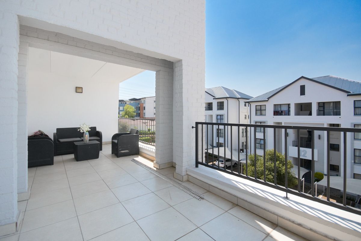 A spacious covered balcony featuring tiled flooring, white painted brick walls, and a metal balustrade overlooking the apartment complex grounds.