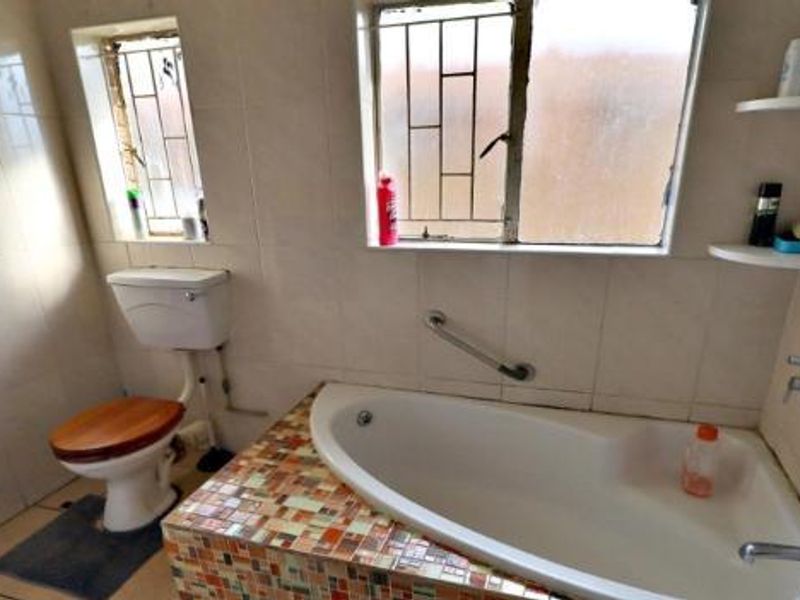 A functional bathroom featuring a built-in bathtub with mosaic tile surround, ceramic wall and floor tiling, and a fitted toilet with security bars on windows.