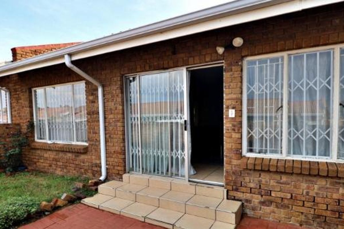 Rear exterior view of a face brick townhouse featuring a sliding glass door with security gates, tiled entrance steps, and a small garden area.