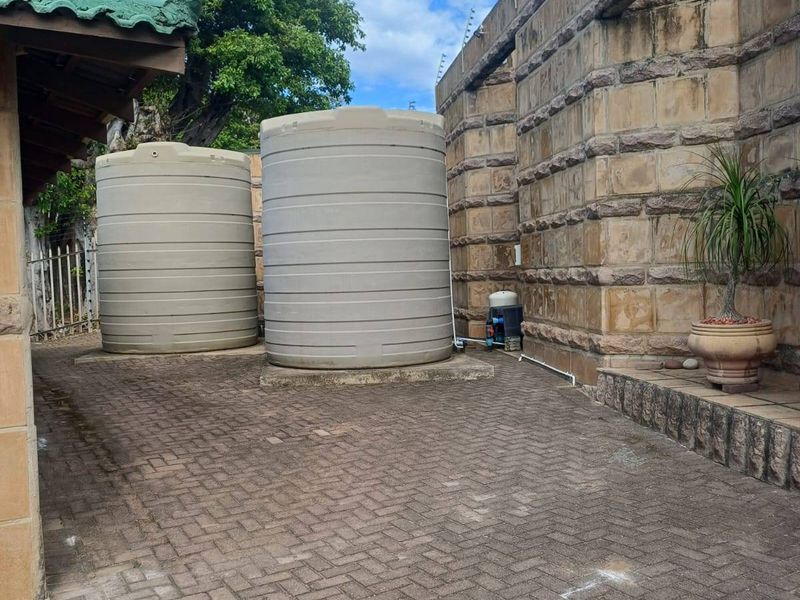 A paved rear courtyard area featuring two large water storage tanks, a water pump, and a high stone boundary wall with electric fencing.