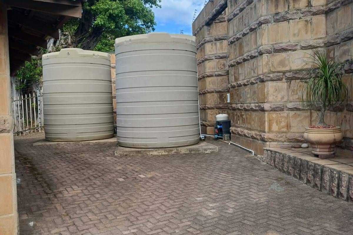 A paved rear courtyard area featuring two large water storage tanks, a water pump, and a high stone boundary wall with electric fencing.