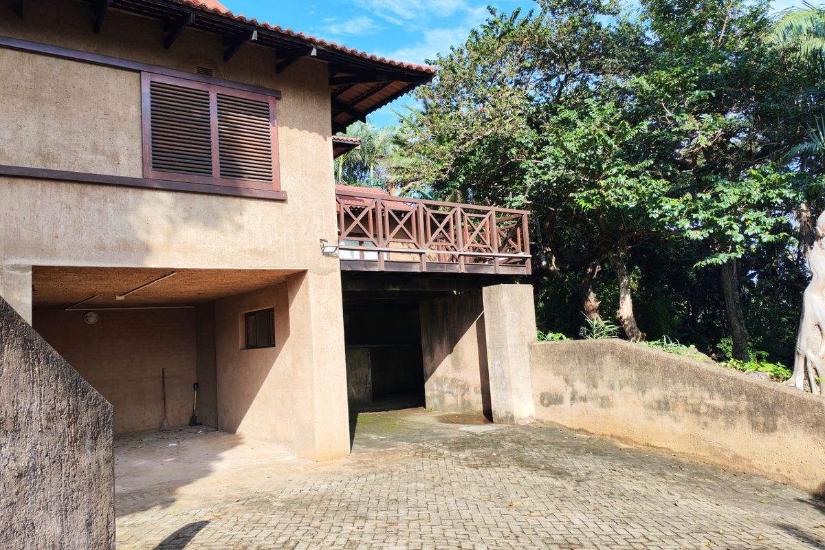 Rear exterior view of the property featuring a paved driveway, a timber balcony, and a retaining wall set against a lush garden backdrop.