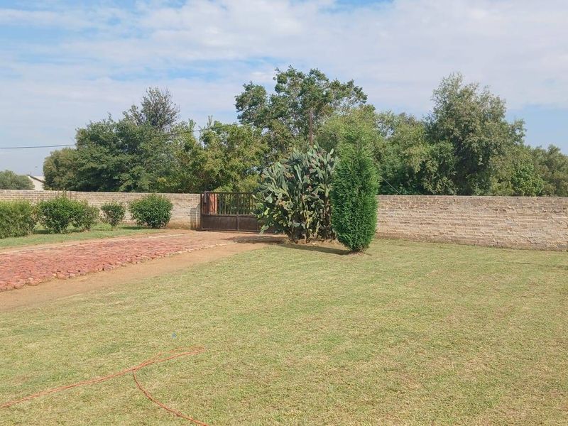 A spacious garden area featuring a well-maintained lawn, a brick-paved driveway, and a secure perimeter boundary wall with a metal entrance gate.