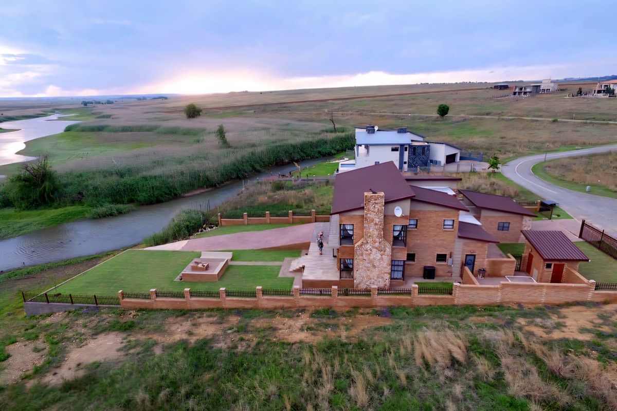 Aerial view of a brick house with a stone chimney, metal roof, and landscaped garden overlooking a river and open landscape.