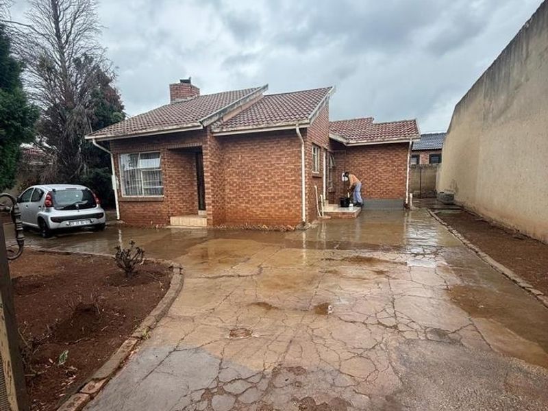 Front exterior view of a single-story brick house featuring a tiled roof, chimney, and a large concrete driveway in need of repair.