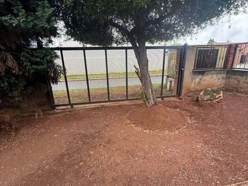 Front garden area featuring a metal driveway gate, perimeter wall, and brick edging, with a mature tree as a central focal point.