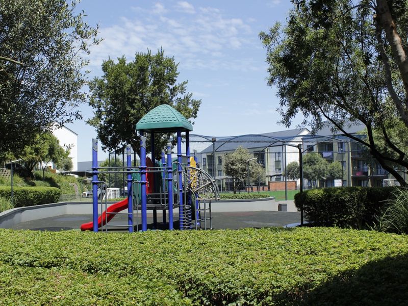 A well-maintained communal garden area featuring a playground, landscaped hedges, and secure fencing within the apartment complex grounds.
