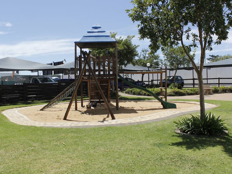 A well-maintained communal garden area featuring a children's playground, sand pit, manicured lawn, and shaded parking structures in the background.