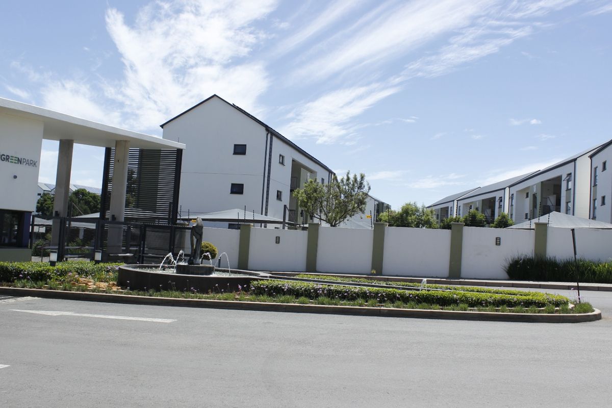 The entrance to Green Park apartment complex featuring a modern security gatehouse, perimeter wall with electric fencing, and a landscaped water fountain feature.