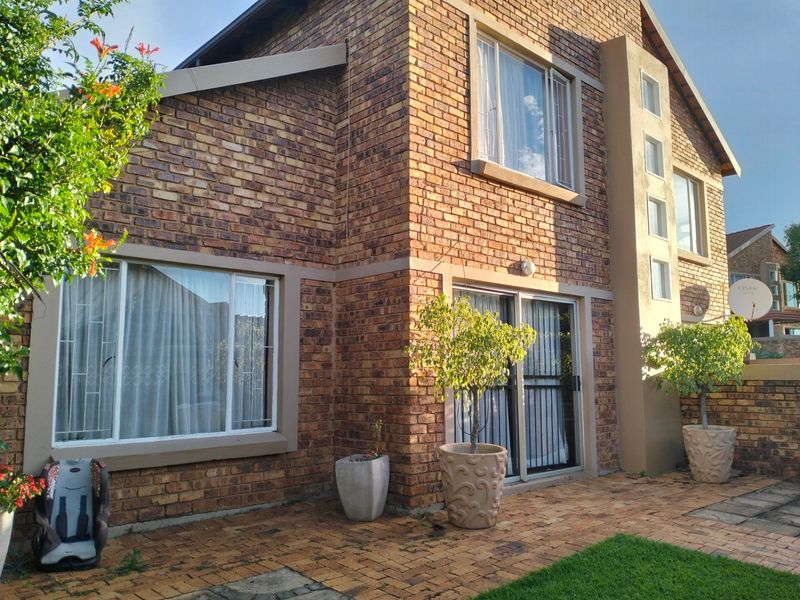 Rear view of a double-storey face brick townhouse featuring a paved patio area, sliding glass doors, and established garden elements.