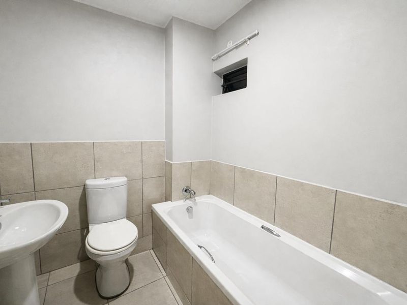 A clean, modern bathroom featuring a white pedestal basin, toilet, and bathtub with neutral ceramic tiling throughout.