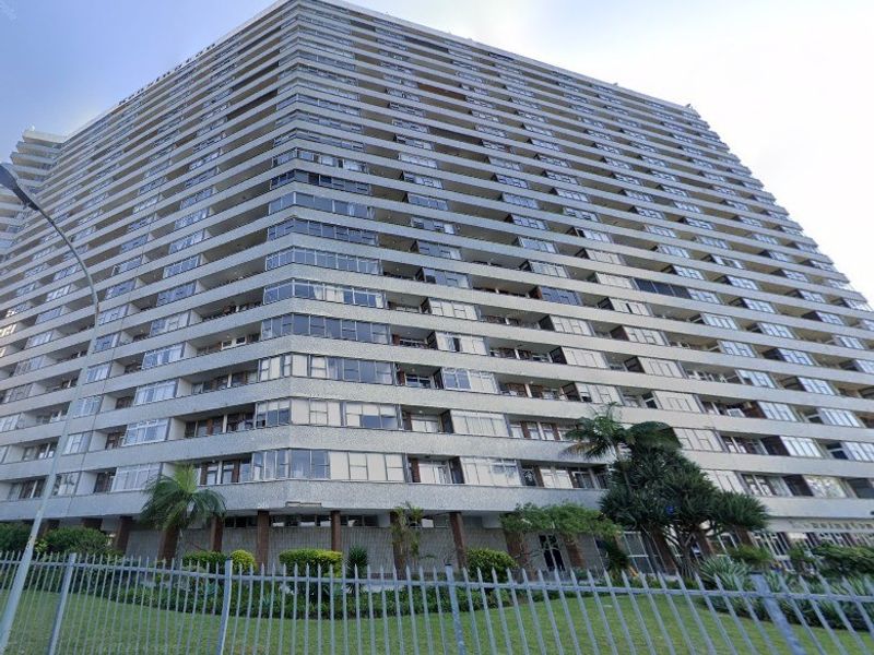 A low-angle exterior view of a large, multi-story apartment building featuring numerous balconies and a fenced garden area at the base.