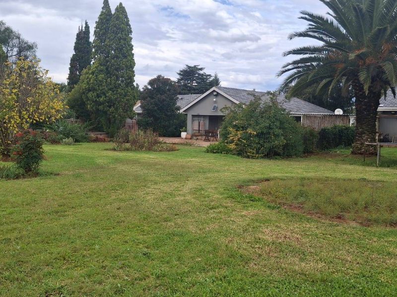 A spacious rear garden featuring a well-maintained lawn, mature trees, and a paved patio area attached to the main house structure.