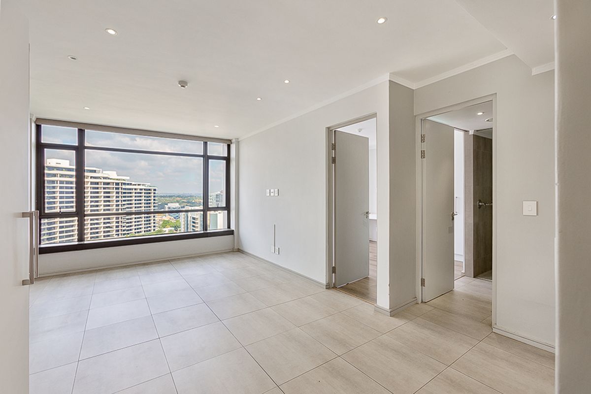 A bright, empty living area featuring large floor-to-ceiling windows with city views, neutral tiled flooring, and recessed lighting.
