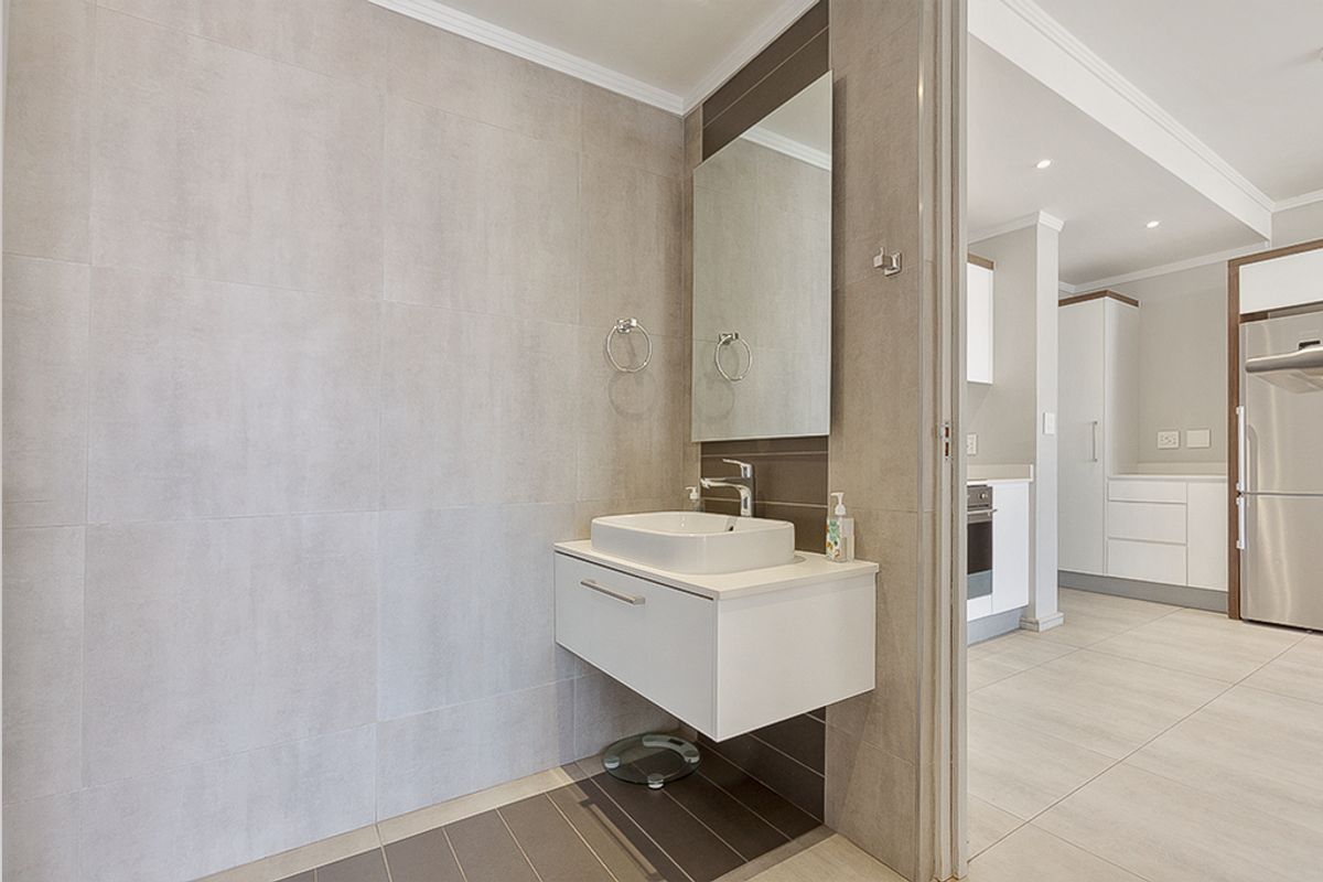 A modern, well-maintained bathroom featuring a wall-mounted vanity with a vessel sink, neutral tiled walls, and a sleek mirror, adjacent to the kitchen area.