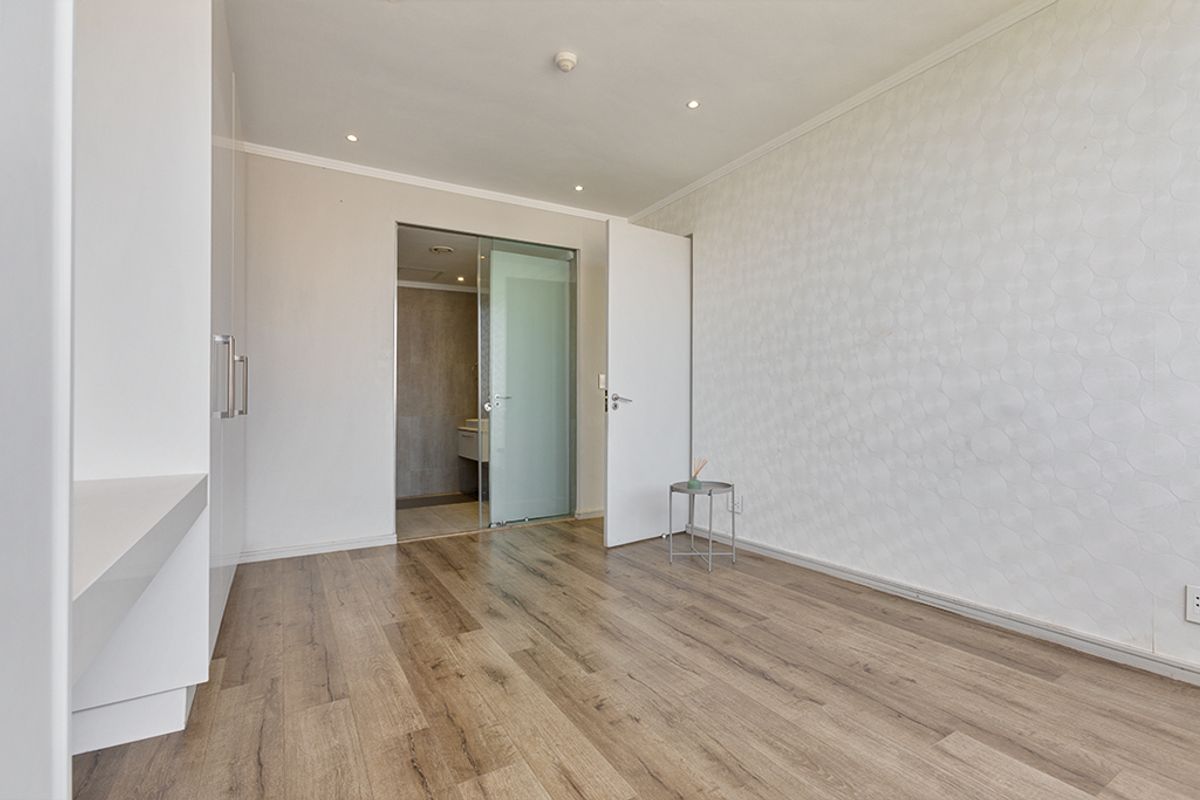 A bright, empty bedroom featuring modern laminate flooring, built-in cupboards, and a glass sliding door leading to an en-suite bathroom.