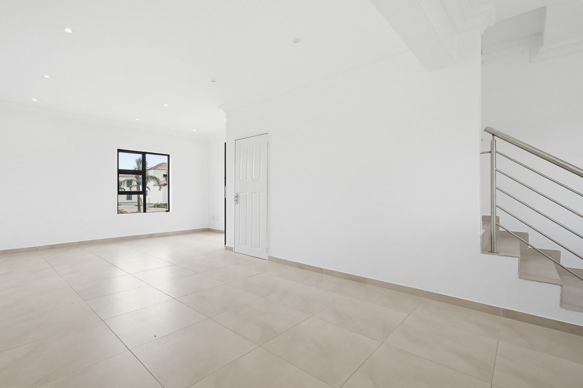 A bright, open-plan living area featuring neutral tiled flooring, recessed lighting, and a modern staircase with a stainless steel balustrade.