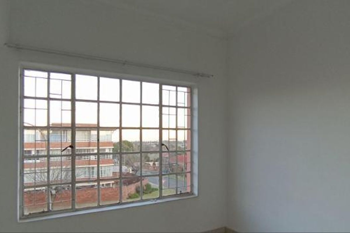 A vacant bedroom featuring a large window with security bars and a curtain rail, offering a view of the surrounding neighborhood.